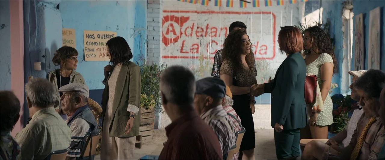 A Hipster in Rural Spain (2024) movie still: medium — A rustic community space features textured blue walls and painted slogans on brick. Two women…