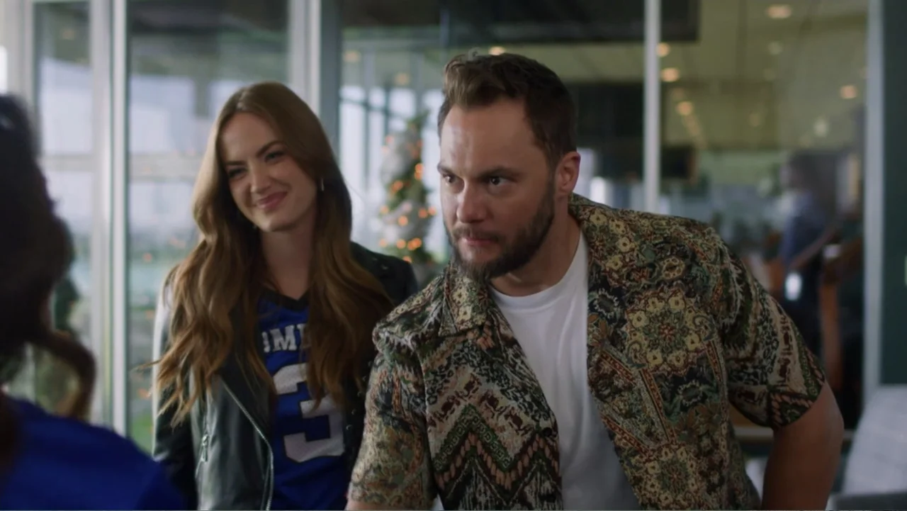 Christmas in the Spotlight (2024) movie still: medium — A modern building lobby with glass partitions and festive decor in the distance. A man and a…