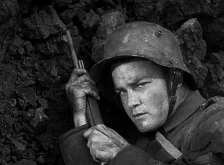 All Quiet on the Western Front (1930) movie still: closeup — A soldier in a metal helmet crouches within a narrow, dirt-walled trench. He holds a…