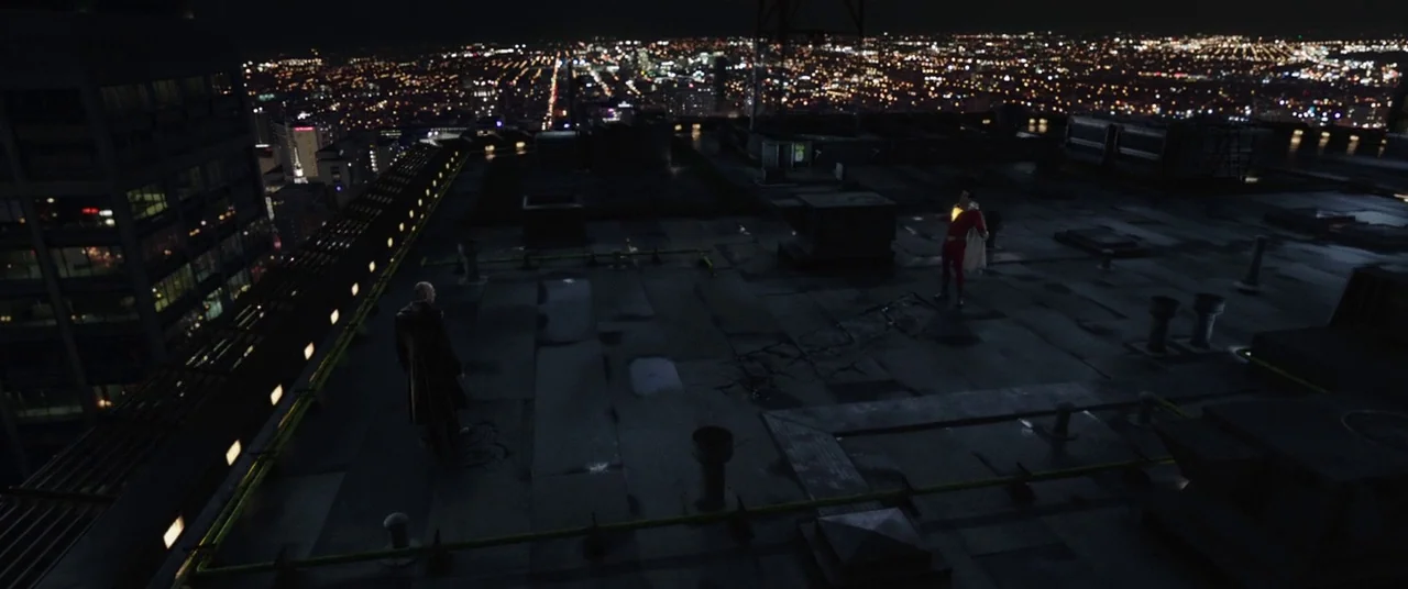 Shazam! (2019) movie still: wide — A dark, flat industrial building roof overlooks a sprawling city grid at night. One man stands in a trench coat…