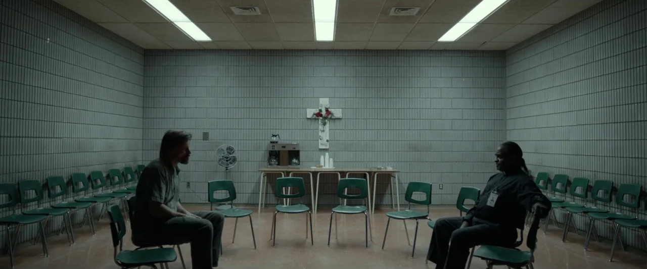 Small Crimes (2017) movie still: wide — A square room with cinderblock walls and rows of green plastic chairs arranged circular. Two men sit…