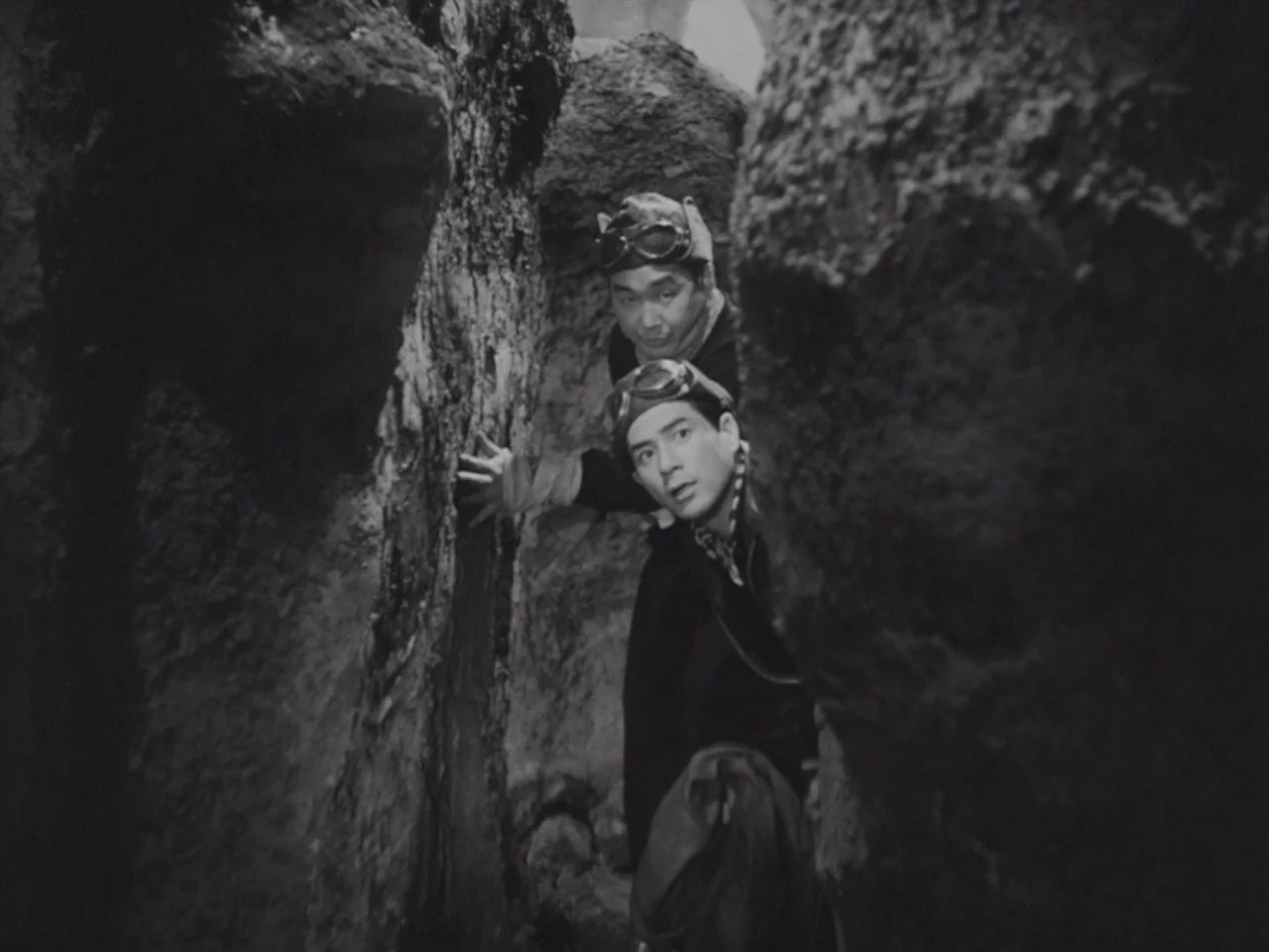 Godzilla Raids Again (1955) movie still: medium — Two men traverse a narrow path between jagged rock formations in an outdoor setting. The men wear…