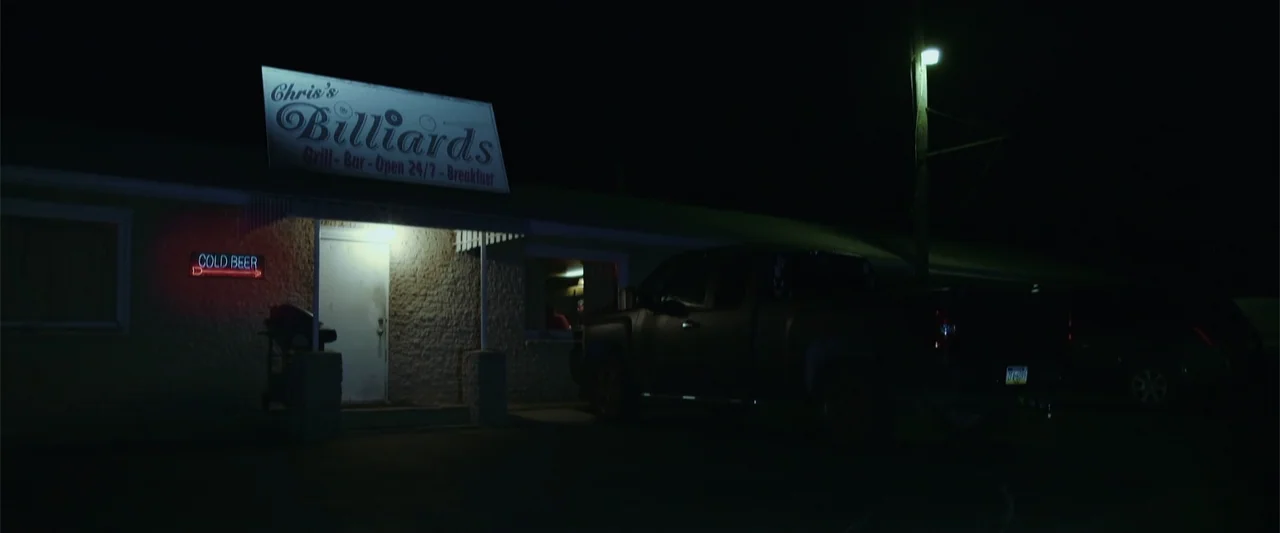Darkness Waits (2020) movie still: wide — A single-story commercial building exterior at night features a prominent sign for a billiards hall. A…