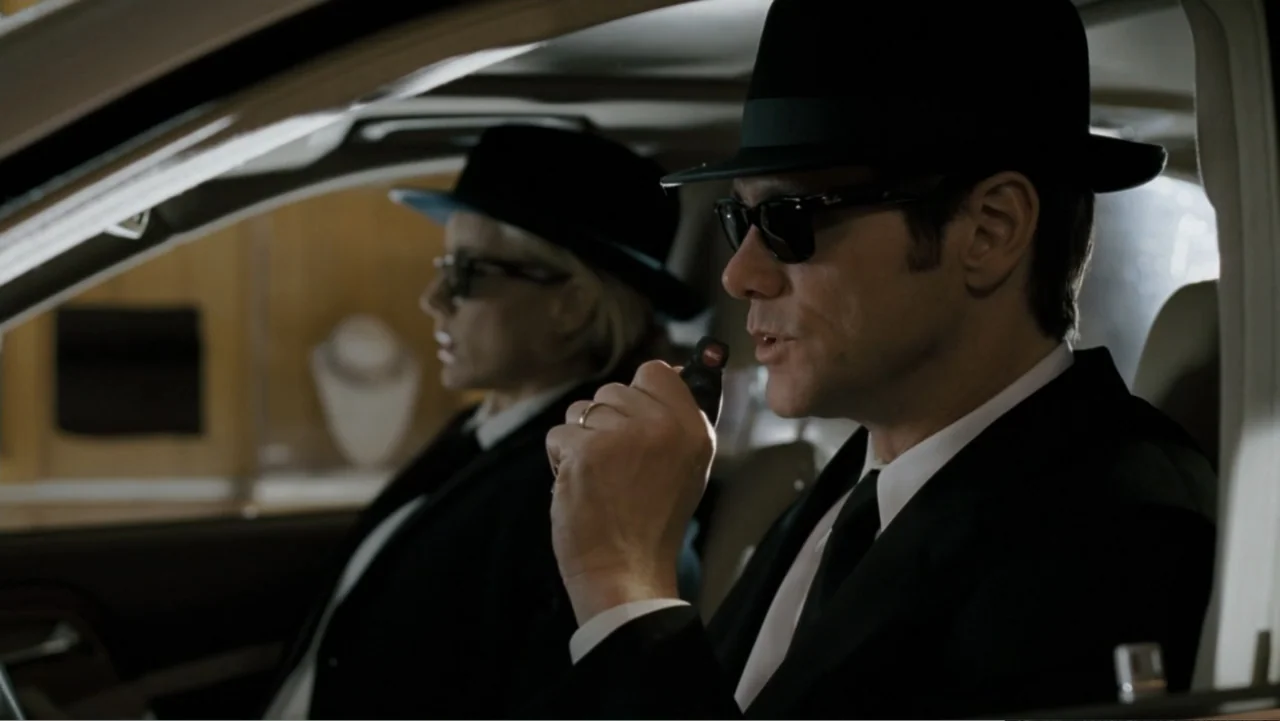 Fun with Dick and Jane (2005) movie still: medium — Two people in suits sit inside a vehicle during the day. The man in the foreground speaks while…