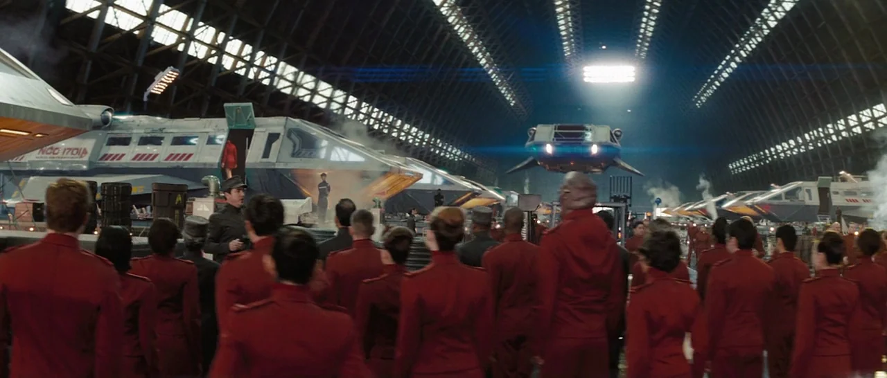 Star Trek (2009) movie still: wide — A vast, vaulted metal hangar contains multiple spacecraft parked in rows under bright lights. A large crowd of…