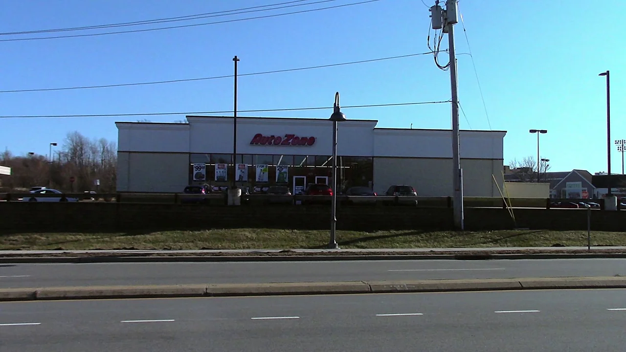 We’re All Going to the World’s Fair (2021) movie still: establishing — An AutoZone retail store is situated behind a grassy embankment and a paved…