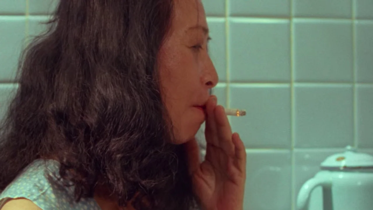 Shirkers (2018) movie still: closeup — A woman stands in a kitchen with seafoam green square tiles on the wall. She has dark hair and holds a lit…