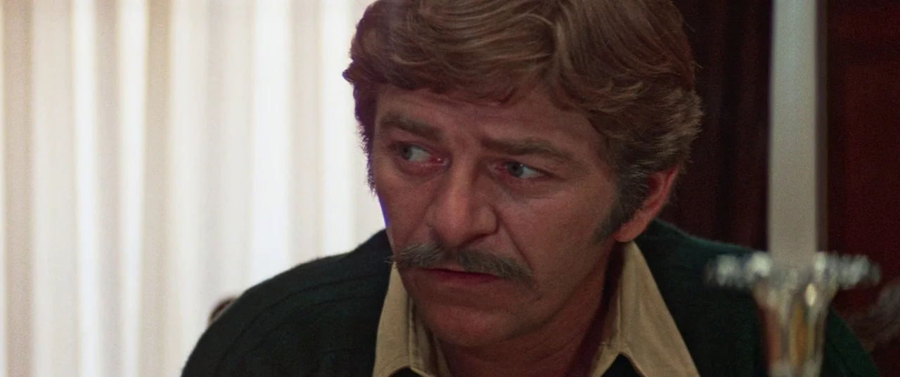 Death Game (1977) movie still: closeup — A man sits inside a room with vertical blinds visible in the background. The man looks toward the side of…
