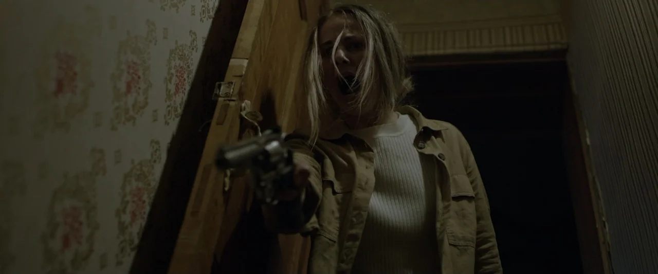 Motherly (2022) movie still: medium — A dimly lit narrow hallway with floral patterned wallpaper features wood paneled details. A woman stands in…