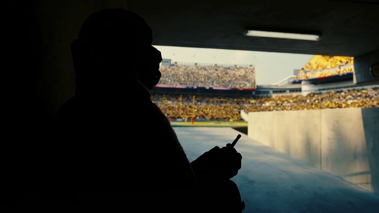 The Dark Knight Rises (2012) movie still: medium — A dark concrete tunnel leads out into a bright, crowded sports arena with yellow seats. A…