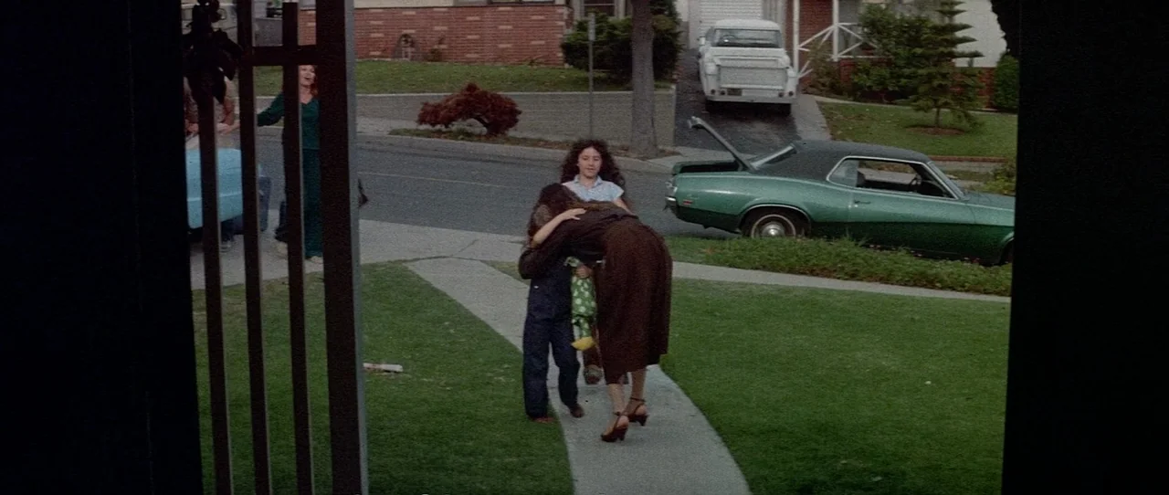 The Entity (1982) movie still: fullbody — A suburban front yard features a concrete path, green lawn, and parked vintage vehicles. A woman in a…