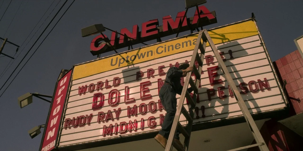 Dolemite Is My Name movie still: fullbody — A tall outdoor theater marquee with red and yellow signage stands against a clear sky. A man in a blue…