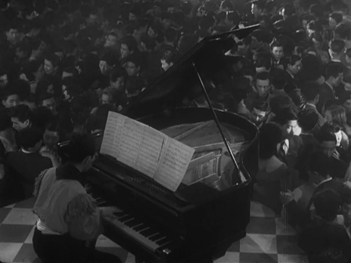 Ikiru (1952) movie still: wide — A crowded dance hall features a polished grand piano on a checkered floor. A man in a decorative jacket plays the…