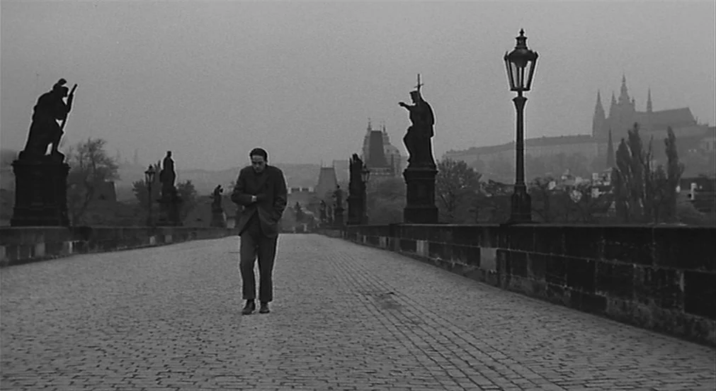 Kafka (1991) movie still: fullbody — A stone paved bridge lined with baroque statues stretches toward a distant gothic cathedral. A man walks toward…
