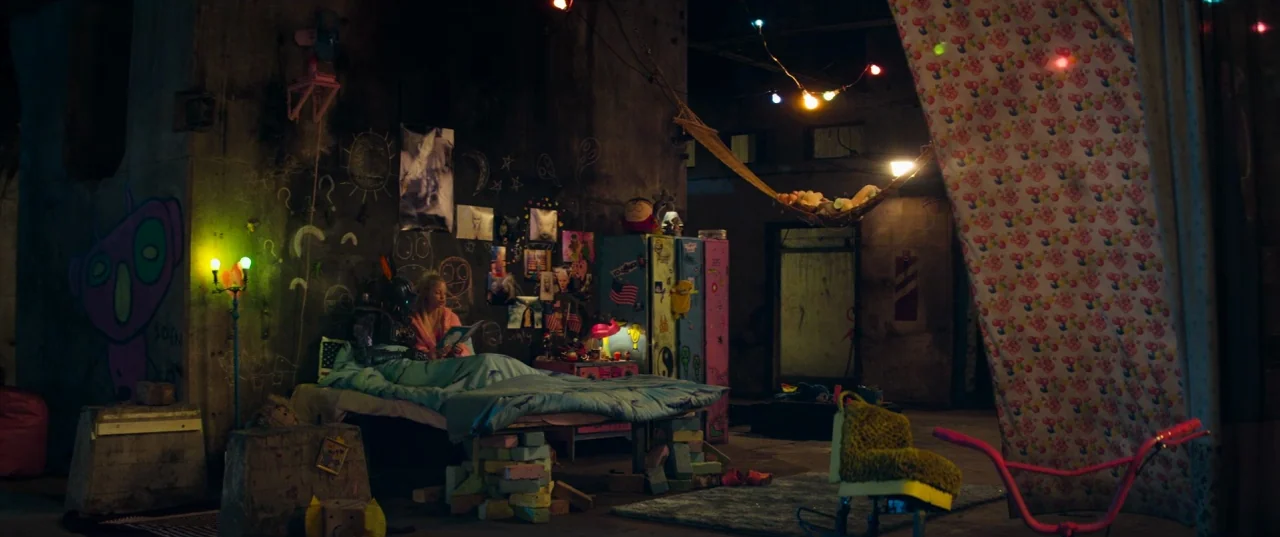 Chappie (2015) movie still: wide — A cluttered industrial space serves as a makeshift home filled with colorful graffiti and toys. A young girl…