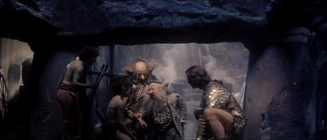 Legend (1985) movie still: medium — A stone cavern interior with rough textured walls and thick plumes of smoke drifting. A group of four fantasy…