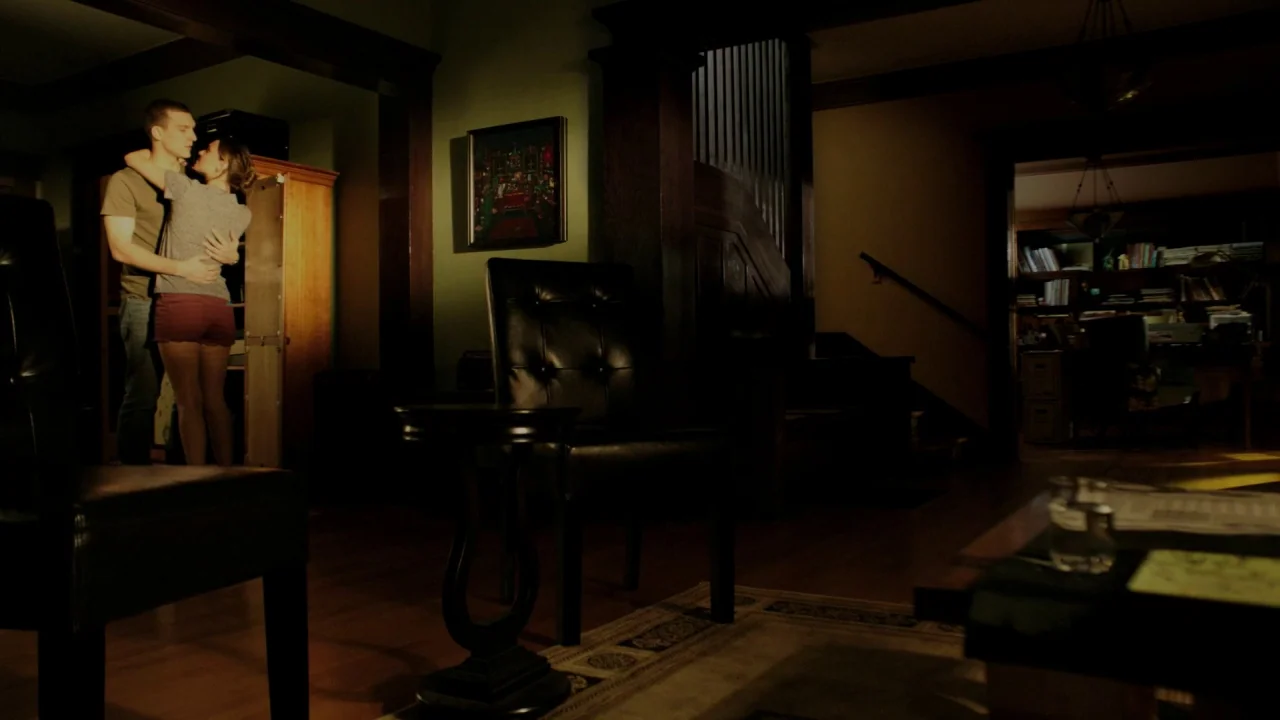 The Pact II movie still: wide — A dimly lit room with dark green walls features a wooden staircase and a bookshelf. A man and a woman stand together…