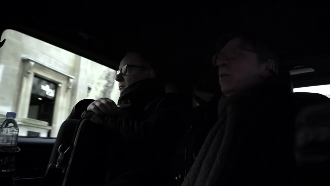 Brexit: Behind Closed Doors (2019) movie still: medium — The interior of a moving car is dark with silhouettes of two men sitting inside. Through…