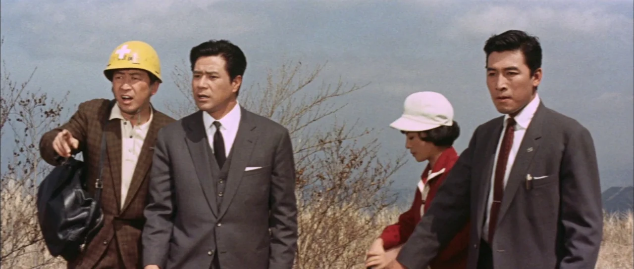 Mothra Vs Godzilla (1964) movie still: medium — A group of four people stands on a dry, brush-covered hillside under a cloudy sky. The subjects look…