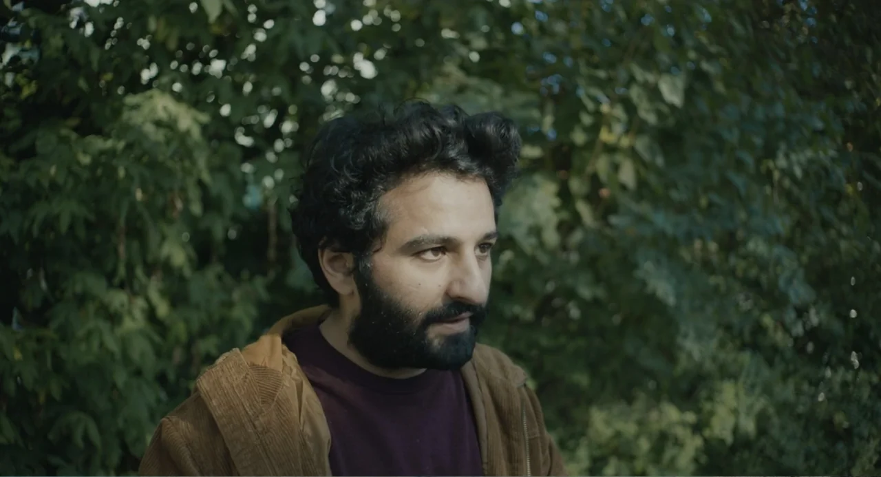 Ninjababy (2021) movie still: medium — A man stands outdoors in front of dense green leafy bushes and trees. He looks to the right side of the frame…