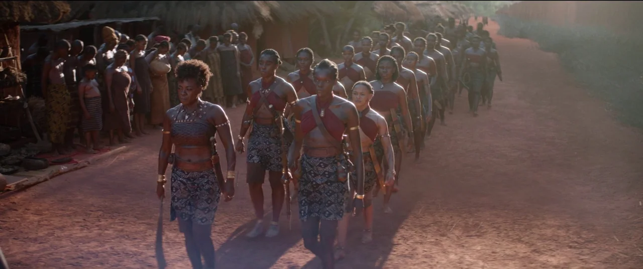 The Woman King (2022) movie still: wide — A group of women in traditional attire marches along a wide, sunlit red-earth path. Thatched-roof huts…