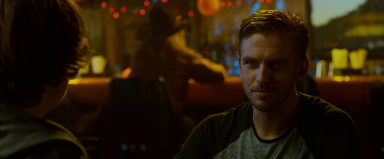The Guest (2014) movie still: medium — A dimly lit restaurant interior features blurred colorful lights in the background. A man sits at a table…
