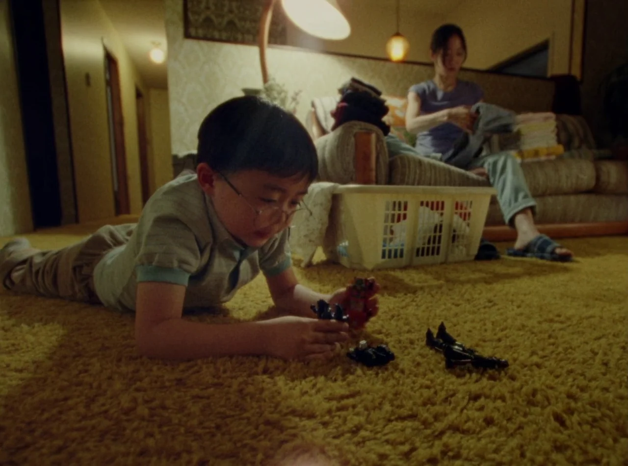 Riceboy Sleeps (2022) movie still: medium — A shag-carpeted room contains a child playing on the floor and a woman nearby. A laundry basket sits…
