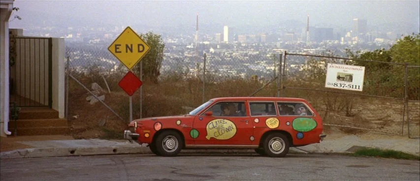 Short Cuts (1993) movie still: wide — A red station wagon decorated with polka dots and text sits near a chain-link fence. The background displays a…