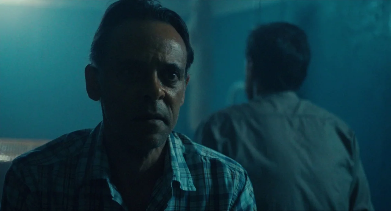 Submergence movie still: medium — A dark indoor space is filled with a dense, teal-tinted atmospheric haze. One man faces the camera directly while…