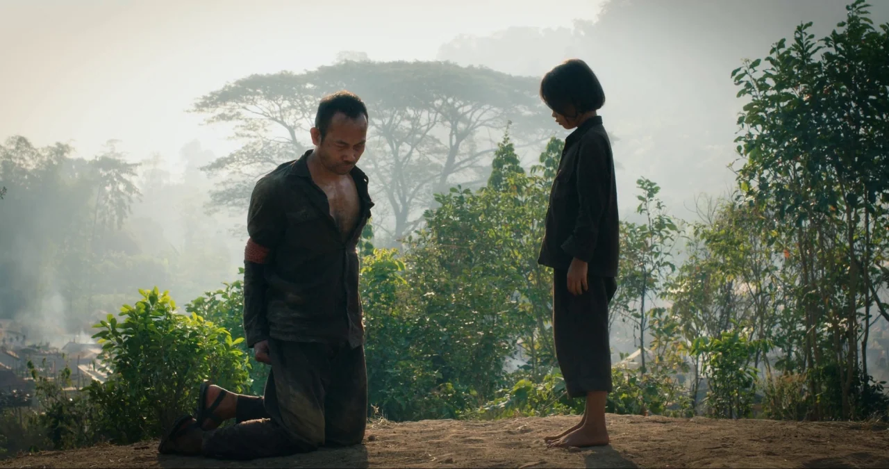 First They Killed My Father (2017) movie still: fullbody — An outdoor clearing features dense foliage and smoke rising from a distant village. A…