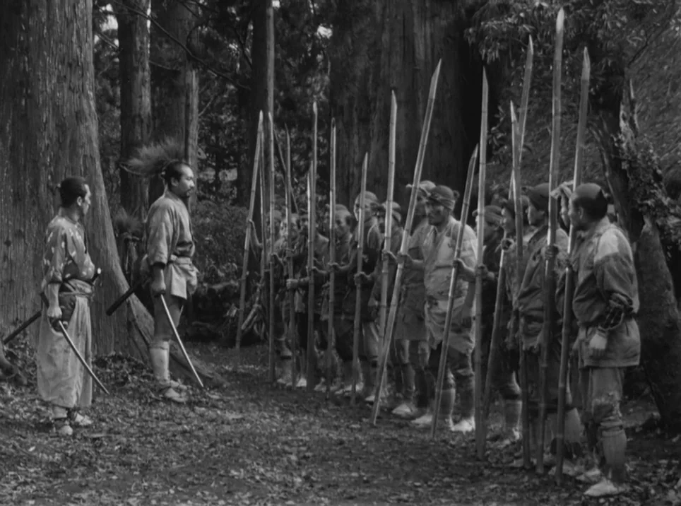 Seven Samurai (1954) movie still: fullbody — A group of men holding long spears stands in a line near large trees. Two men stand opposite the group…