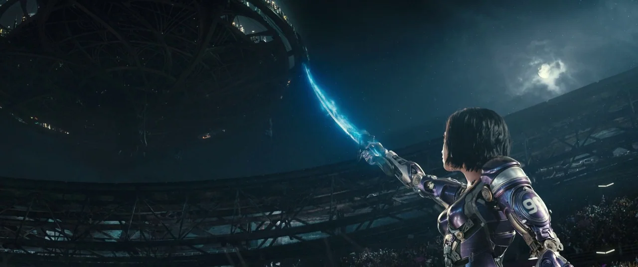 Alita: Battle Angel movie still: wide — A cyborg stands in a vast stadium under a large hovering circular city structure. She points a glowing blue…