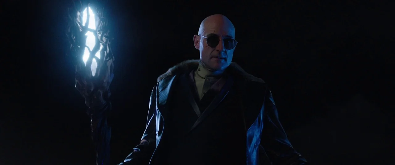 Shazam! (2019) movie still: medium — A bald man stands against a dark, featureless void holding an illuminated staff. The glowing staff illuminates…