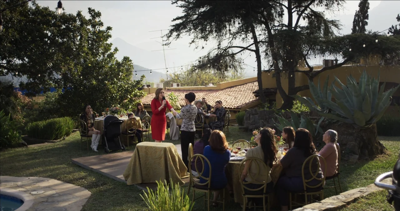 Bendita Suegra (2023) movie still: wide — A festive outdoor gathering takes place on a lawn during the late afternoon. One woman in a red dress…