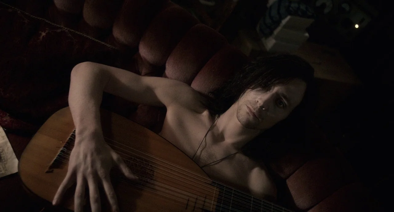 Only Lovers Left Alive (2013) movie still: medium — A dark space features a deep red upholstered sofa with tufted buttons. A bare-chested man with…