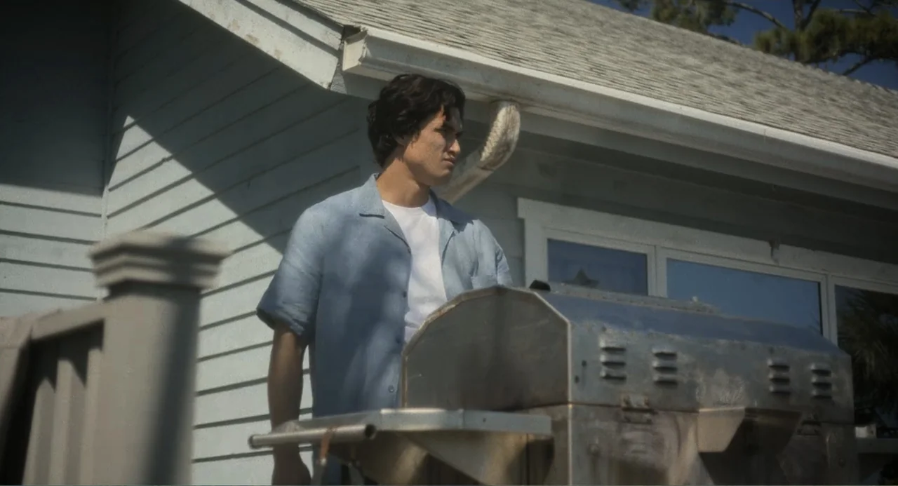 May December (2023) movie still: medium — A light blue-sided house with a wooden deck and a stainless steel grill. A young man stands on the deck…