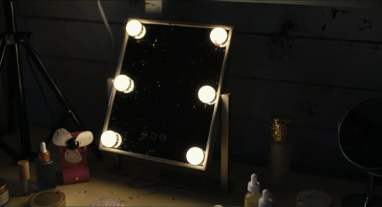 Hell of a Summer (2025) movie still: medium — A dark dressing table holds a lit vanity mirror surrounded by various cosmetic bottles. A small desk…