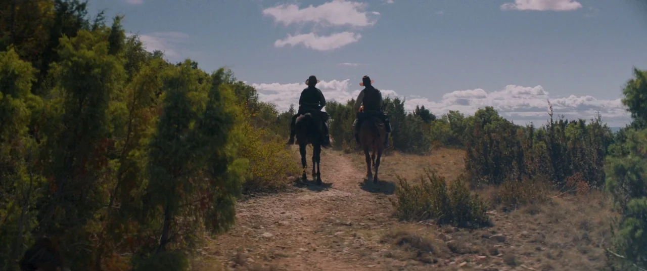 The Sisters Brothers (2018) movie still: wide — A dry dirt trail winds through a sparse landscape covered in low green bushes. Two men on horseback…