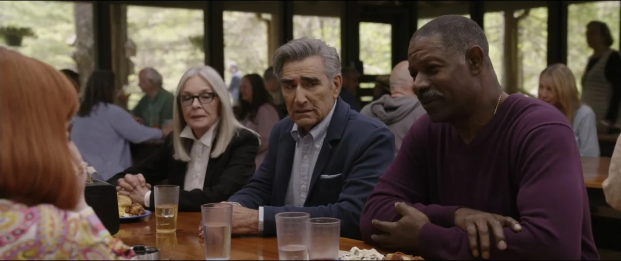Summer Camp (2024) movie still: medium — A group of people sits at a wooden table inside a lodge-style dining hall. Three central figures converse…
