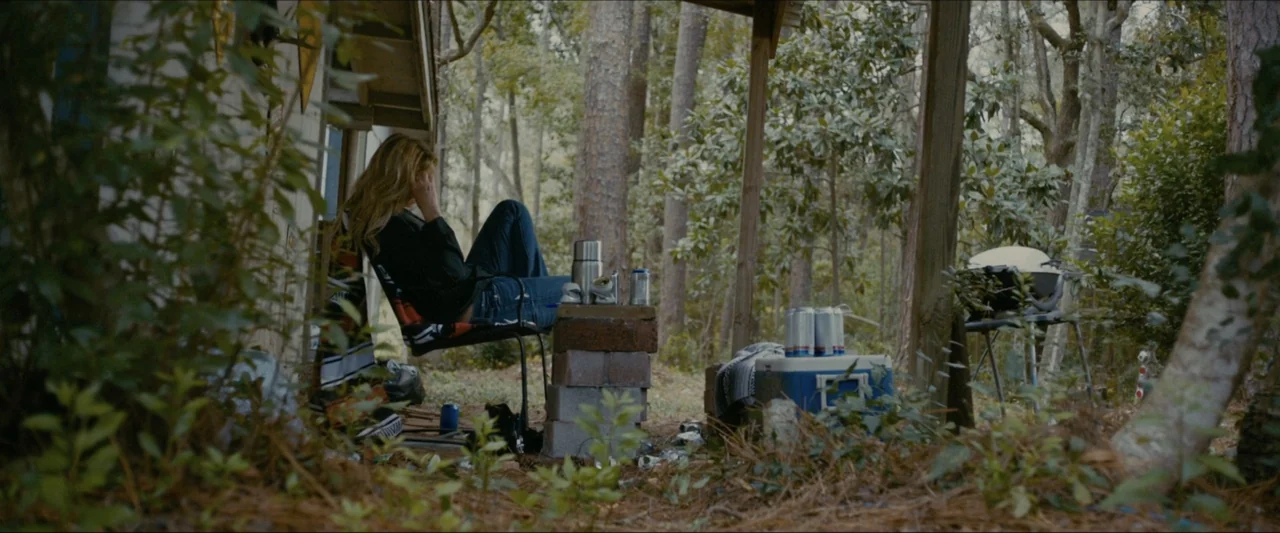 Ambush (2020) movie still: medium — A wooden porch sits at the edge of a dense, shaded pine forest. A woman sits in a chair with aluminum cans on a…
