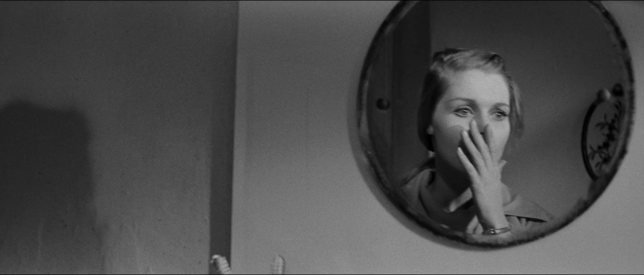Bunny Lake Is Missing (1965) movie still: closeup — A dark bathroom setting features a circular mirror mounted on a plain wall. A woman looks at her…