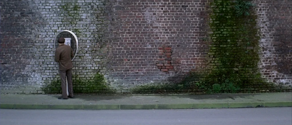 The Banishment (2007) movie still: wide — A weathered brick wall covered in patchy moss runs parallel to a paved sidewalk. A man stands at a…