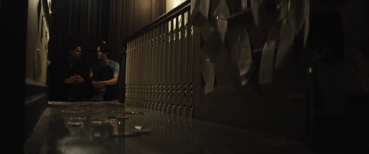 Daniel Isn’t Real movie still: wide — A dark hallway at night has numerous white papers taped to the wall surfaces. Two men sit on the glossy floor…