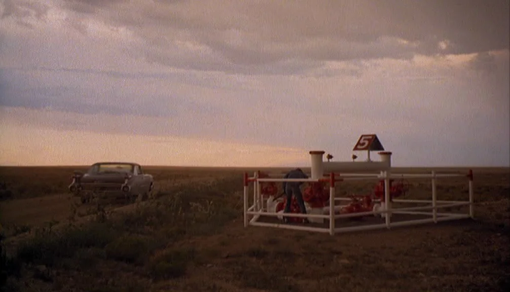 Badlands (1973) movie still: wide — An open, flat expanse of desert terrain under a clouded sunset sky. A person works at a fenced oil wellhead…