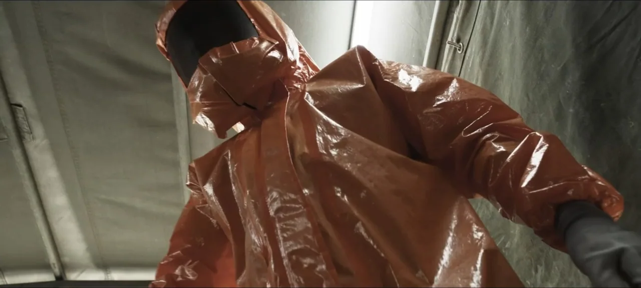 Containment (2015) movie still: medium — An interior space enclosed by grey fabric walls appears to be a medical tent. A figure in a reflective…