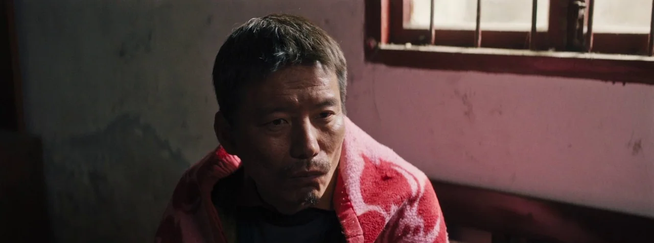 Dead Pigs (2018) movie still: medium — A plain interior room with a window featuring dark metal bars on the wall. A man sits while wearing a bright…
