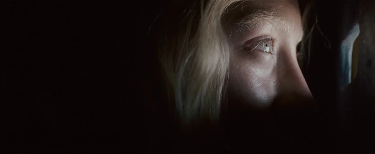 Hanna (2011) movie still: closeup — A dark, enclosed space with only a sliver of light revealing the subject. A young woman with blonde hair stares…