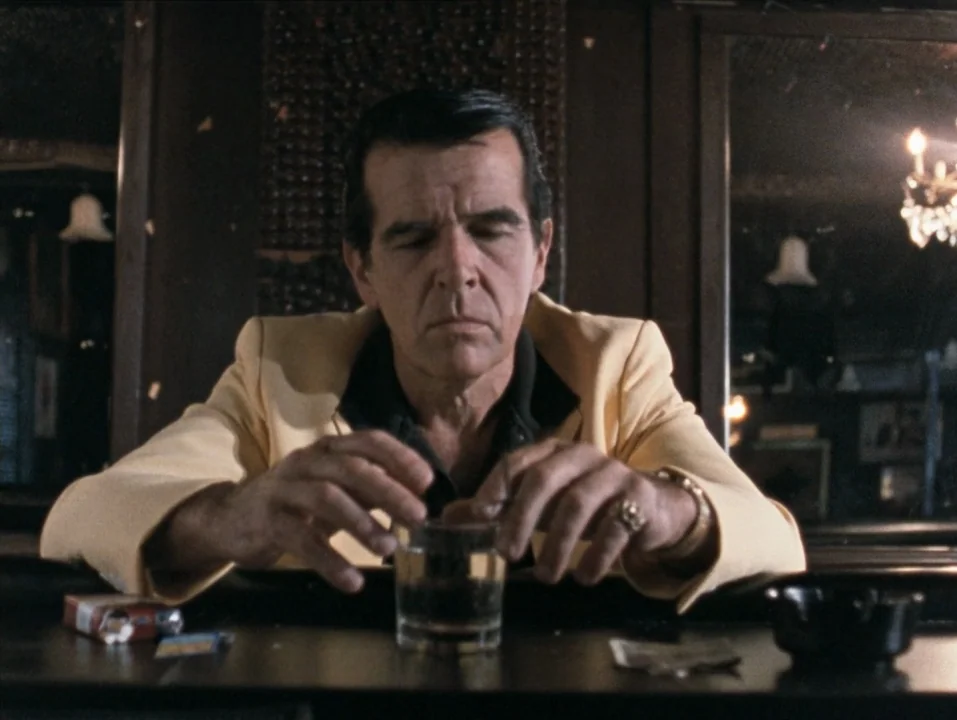 River of Grass (1994) movie still: medium — A man in a yellow blazer sits alone at a dark bar counter. He stares down at his glass with his hands…