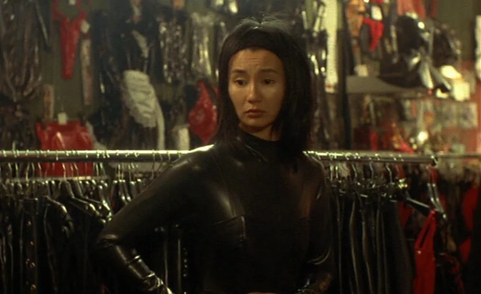 Irma Vep (1996) movie still: medium — A clothing store rack filled with black leather and red vinyl garments fills the space. A young woman in a…