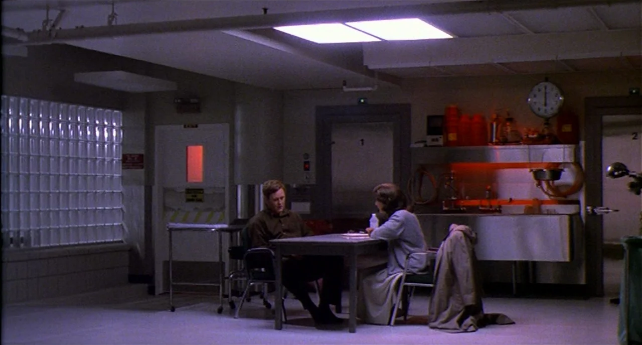 Raising Cain movie still: wide — A utilitarian laboratory interior features metal cabinets, glass block windows, and a wall clock. Two people sit at…